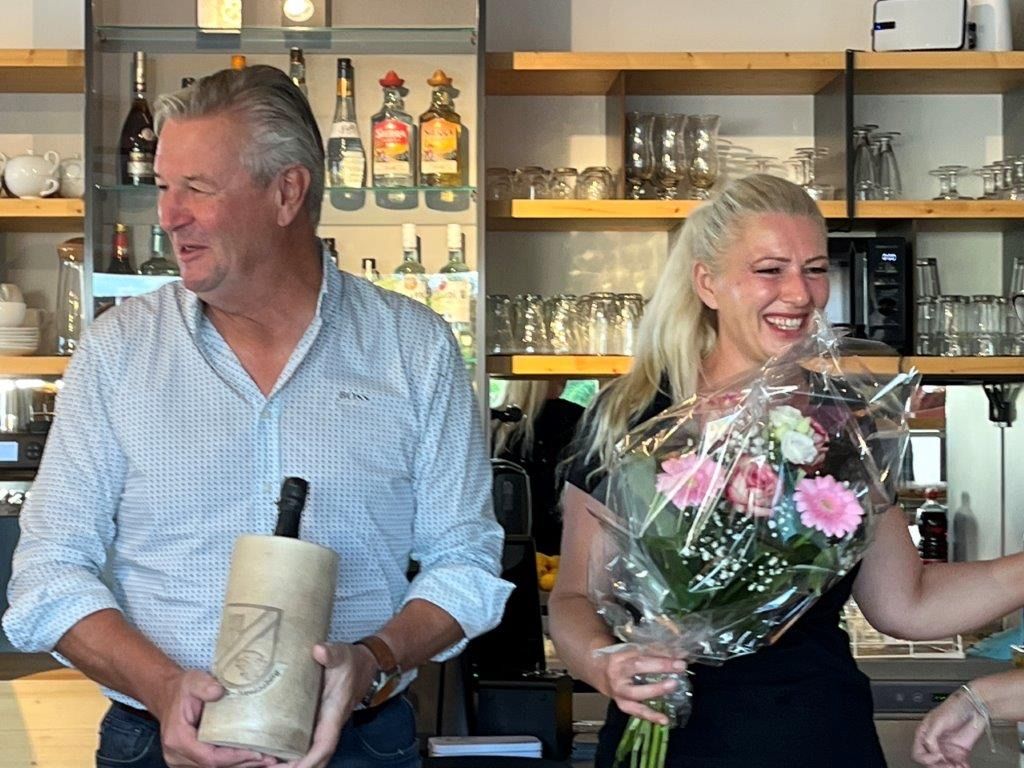 A man holding a bottle and a woman holding a bouquet of flowers stand in a bar.
