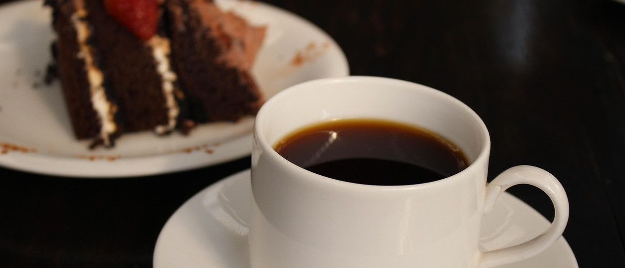 Eine weiße Tasse und Untertasse mit einer vollen Tasse dunklen Kaffee neben einem Teller mit einem Stück Schokoladenkuchen, gekrönt von einer Erdbeere.