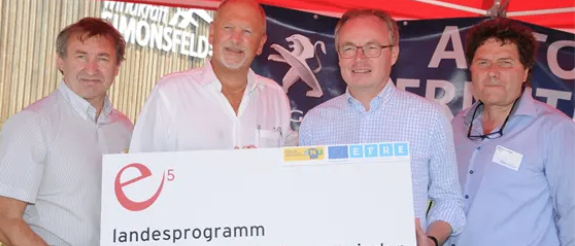 Vier Männer stehen unter einem roten Zelt. Sie halten einen großen Scheck für das Programm energieeffizienter Gemeinden.