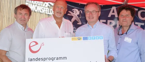 Vier Männer stehen unter einem roten Zelt. Sie halten einen großen Scheck für das Programm energieeffizienter Gemeinden.