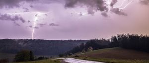 Ein Gewitter mit Blitzen über einer ländlichen Landschaft, mit einem gelben Haus auf einem Hügel und einer Straße, die durch ein Feld führt.