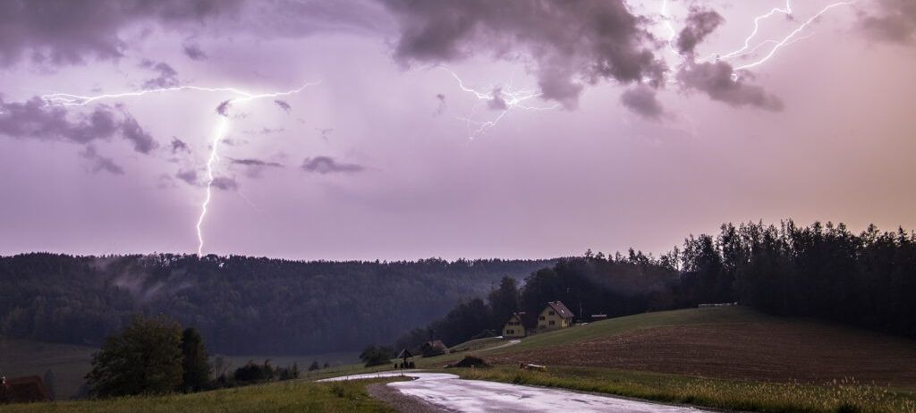 Ein Gewitter mit Blitzen über einer ländlichen Landschaft, mit einem gelben Haus auf einem Hügel und einer Straße, die durch ein Feld führt.