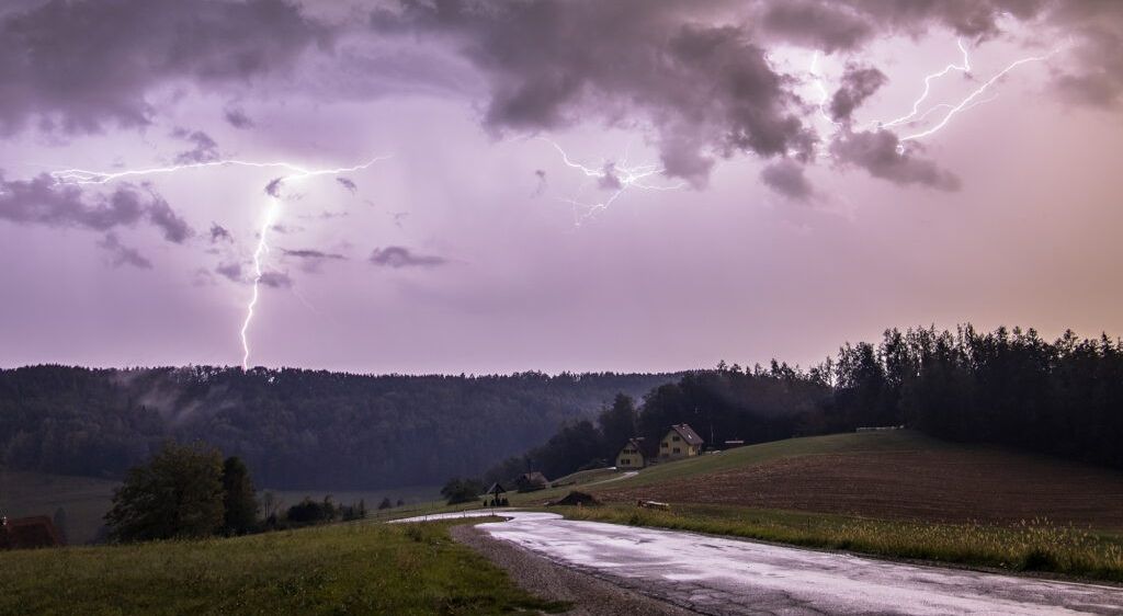 Ein Gewitter mit Blitzen über einer ländlichen Landschaft, mit einem gelben Haus auf einem Hügel und einer Straße, die durch ein Feld führt.