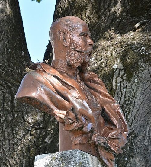 Eine Bronzebüste eines bärtigen Mannes in Uniform steht auf einem Steinsockel unter einem Baum.