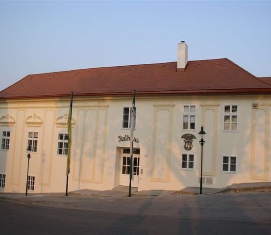 Ein Gebäude mit einem roten Dach und weißen Wänden. Es hat mehrere Fenster und einen Schornstein. Der Name 'Rath us' ist an der Wand. Vor dem Gebäude stehen zwei Fahnen.