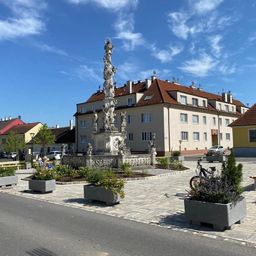 Ein Stadtplatz mit einem Brunnen mit Skulpturen, umgeben von Gebäuden, mit einem Fahrrad und Pflanzen an der Seite.