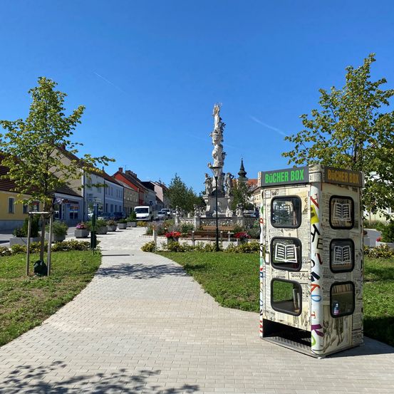 Ein gepflasterter Weg führt zu einer dekorierten Buchbox am Straßenrand mit Gebäuden und einer Statue im Hintergrund.