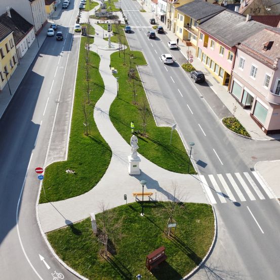 Eine Vogelperspektive einer Straße mit fahrenden Autos. Eine Statue steht in der Mitte der Straße mit grünem Gras auf beiden Seiten. Gebäude in verschiedenen Farben säumen die Straße auf beiden Seiten.