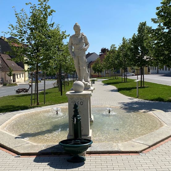 Eine Statue eines Mannes steht in der Mitte eines Brunnens in einem Park, umgeben von Bäumen und Gebäuden. Die Statue steht auf einem Sockel mit einer Kugel, und der Brunnen hat Wasser, das aus ihm fließt.