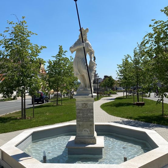 Eine Statue einer Frau mit einer Lanze steht neben einem Brunnen, umgeben von Bäumen und Gras. Der Brunnen ist mit Wasser gefüllt, und die Schatten der Bäume fallen auf das Gras.