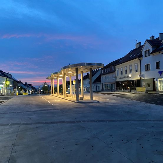 Eine ruhige Straße in der Dämmerung mit einer Tankstelle und Gebäuden auf der rechten Seite. Der Himmel ist blau mit einem Hauch von Rosa.