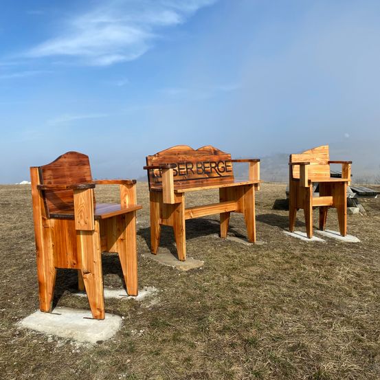 Drei Holzbänke stehen auf einer grasigen Anhöhe unter einem blauen Himmel. Die mittlere Bank hat 'Ober Berge' auf der Rückenlehne geschrieben.