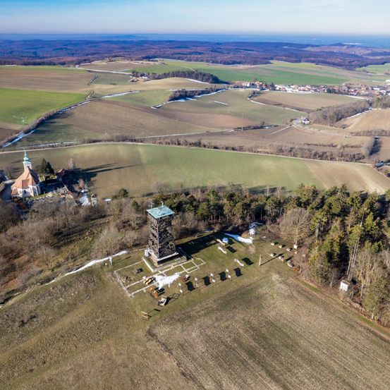 Luftaufnahme einer ländlichen Landschaft mit einem Turm, einer Kirche und Ackerland. Der Turm ist von einer Rasenfläche umgeben und hat ein grünes Dach. Die Kirche befindet sich auf der linken Seite.