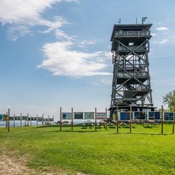 Ein hoher Aussichtsturm steht auf einem Rasenfeld mit blauem Himmel und Wolken. Bänke und Tische sind davor aufgestellt, und eine Person steht in der Nähe des Turms.