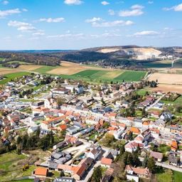 Luftaufnahme einer Stadt mit vielen Gebäuden, umgeben von grünen Feldern und Bäumen unter einem teilweise bewölkten Himmel.