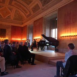 Zwei Musiker spielen auf der Bühne mit Klavier und Cello, während eine Gruppe von Menschen in einem eleganten Raum mit verzierter Decke und Wänden zusieht.