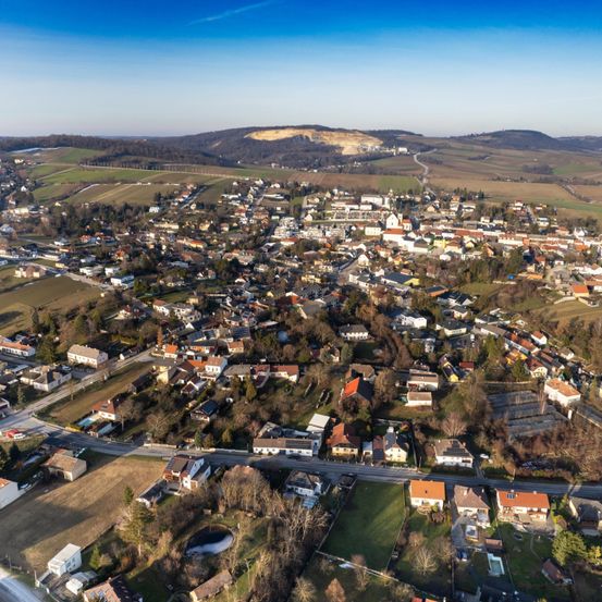 Luftaufnahme einer kleinen Stadt umgeben von grünen Feldern und Hügeln unter einem klaren blauen Himmel.