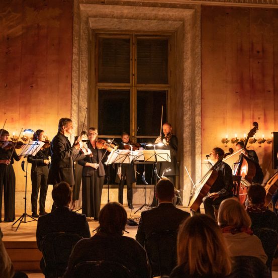 Eine Gruppe von Musikern spielt auf der Bühne vor einem Publikum. Violinen, Violoncelli und andere Instrumente stehen auf Ständern. Die Musiker sind in formeller Kleidung gekleidet. Das Publikum sitzt auf Stühlen. Der Raum hat warmes Licht.