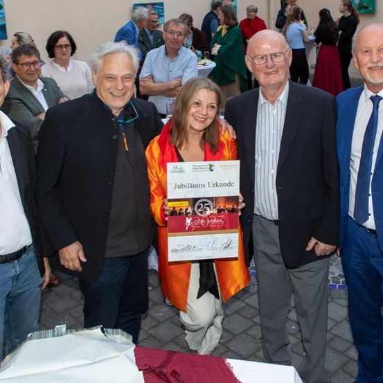 Eine Gruppe von Menschen steht in einer formellen Umgebung zusammen. Eine Frau hält eine Urkunde und lächelt. Hinter ihr stehen drei Männer und eine Frau eng beieinander und lächeln ebenfalls. Sie sind in Anzügen und Krawatten gekleidet, und der Hintergrund ist mit anderen Menschen verschwommen.