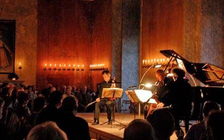 Ein Mann spielt Geige auf der Bühne vor einem Publikum, mit einem Pianisten und einem anderen Geiger. Kerzen sind auf der Bühne angezündet.