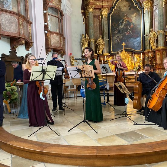 Eine Gruppe von Musikern tritt auf der Bühne in einer Kirche auf, mit Violinen, Celli und einer Flöte. Sie sind in formeller Kleidung gekleidet.