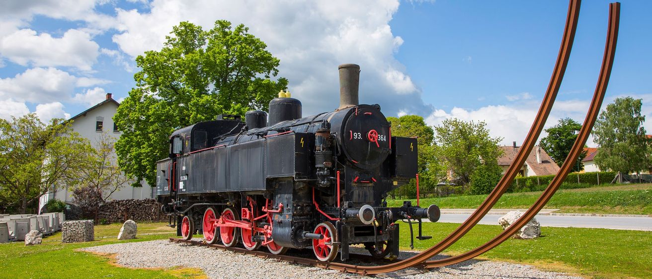 Eine alte schwarze Lokomotive mit roten Rädern steht auf einer Kiesplattform. Sie trägt die Nummer 193 364. Im Hintergrund sind Bäume und ein Haus zu sehen.