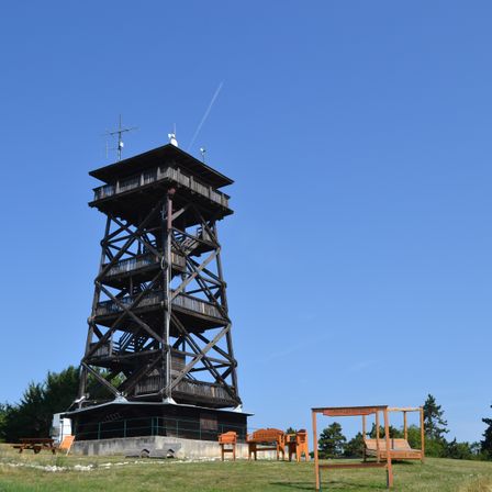 Ein hoher Holzturm steht auf einem grasbewachsenen Hügel bei klarem blauem Himmel. Vor dem Turm sind mehrere orangefarbene Stühle und eine Überdachung aufgestellt. Der Turm hat mehrere Stockwerke und Antennen auf der Spitze.