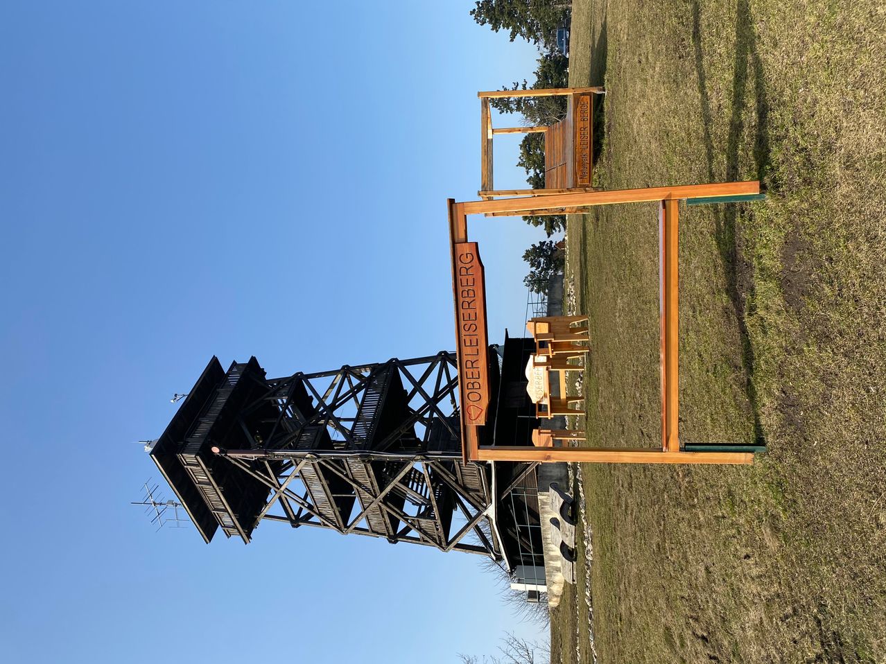 Ein hoher Holzturm namens Oberleerberg steht auf einem Grasfeld unter einem klaren blauen Himmel.