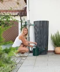 Eine Frau kniet neben einem schwarzen Regentonne und gießt Pflanzen mit einem grünen Gießkannen in einem Garten. Hinter ihr ist ein Holzzaun und ein Haus zu sehen.