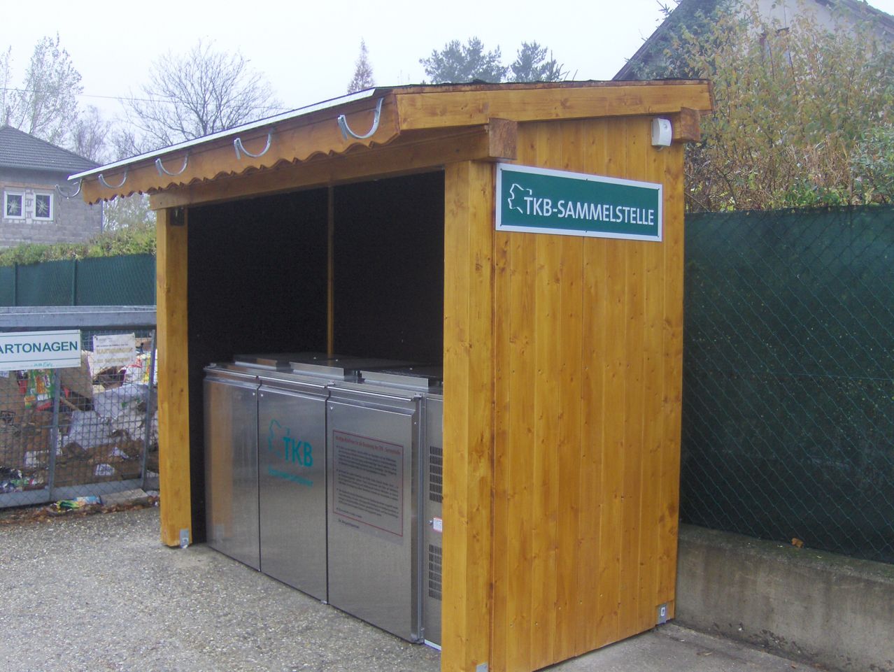 Eine Holzhütte mit einem Metalldach, die einen Metallcontainer mit der Aufschrift 'TKB-Sammelstelle' beherbergt, in einem eingezäunten Bereich mit Bäumen und einem Gebäude im Hintergrund.