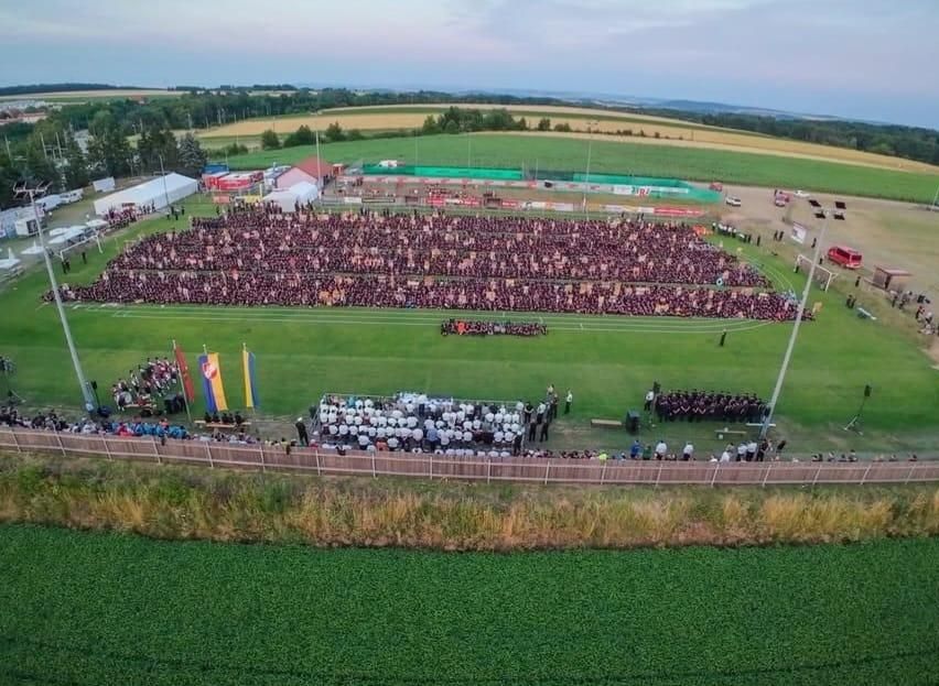 Eine große Menschenmenge versammelt sich auf einem Feld, wahrscheinlich für ein Festival oder eine Veranstaltung. Die Menge sitzt in Reihen auf dem Rasen, und einige Flaggen sind sichtbar.