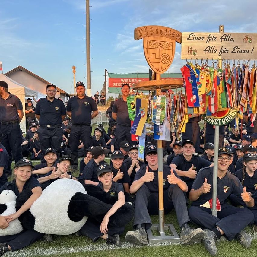 Eine Gruppe von Personen in Uniform posiert für ein Foto, wobei einige Daumen hochhalten, während ein großer Panda-Maskottchen im Vordergrund steht und ein hölzerner Schild mit einer Botschaft dahinter ist.