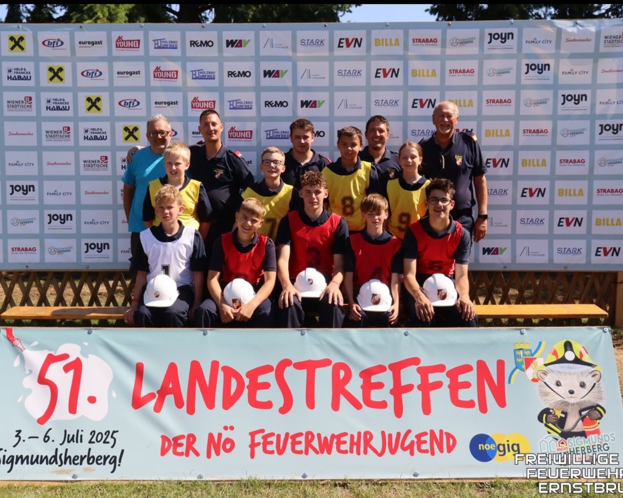 Eine Gruppe junger Menschen in Sportuniformen und weißen Helmen posiert für ein Foto vor einem Banner, das '7. LANDESTREFFEN DER NO FEUWERWEHRJUGEND' besagt. Der Hintergrund zeigt Logos verschiedener Sponsoren.
