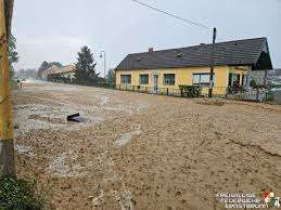 Eine schlammige Straße mit einem gelben Haus im Hintergrund, einer Straßenlaterne und Bäumen. Eine Person steht in der Nähe des Hauses.