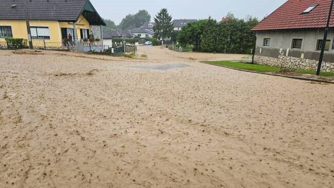 Eine Straße ist mit schlammigem Wasser überflutet. Ein Haus mit gelber Außenseite und grünem Vordach steht links. Ein graues Haus mit rotem Dach steht rechts. Bäume und Büsche sind im Hintergrund.