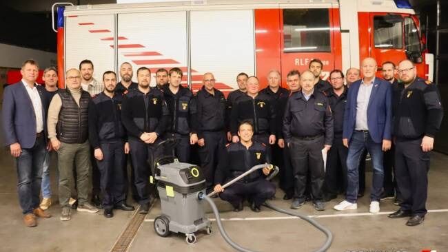 Eine Gruppe von Feuerwehrleuten und Offiziellen posiert für ein Foto mit einem Feuerwehrwagen und einem Staubsauger in einer Garage.