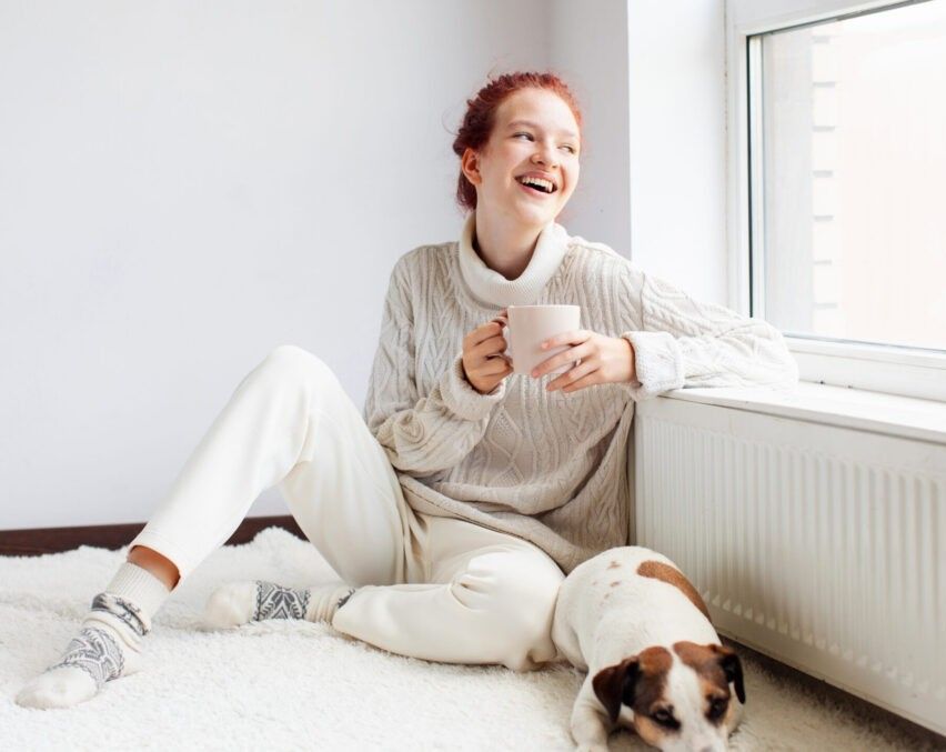 Eine Frau mit roten Haaren lächelt, während sie auf einem weißen Teppich sitzt, einen weißen Becher hält und einen weißen Rollkragenpullover und Hosen trägt. Ein weißer und brauner Hund sitzt neben ihr.