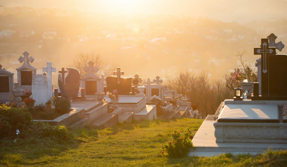 Ein Friedhof mit Grabsteinen und Kreuzen ist vor einem goldenen Sonnenuntergang. Die Gräber sind in Reihen angeordnet, einige mit Blumen. Bäume und Gebäude sind in der Ferne sichtbar.