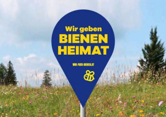 Ein blaues Schild mit dem Text Wir geben Bienen Heimat steht auf einem Pfahl in einem Blumenfeld.