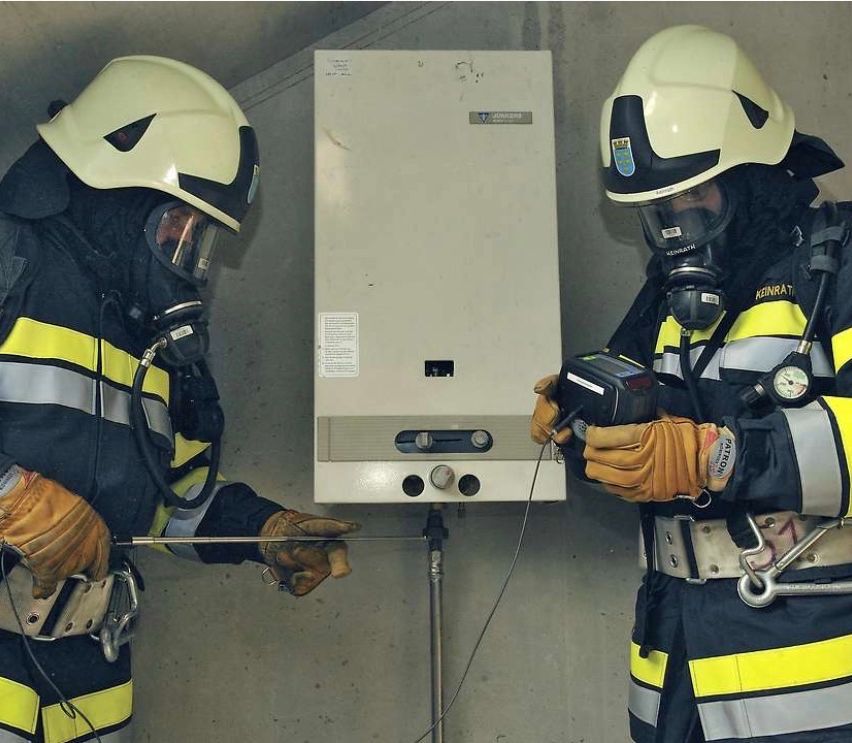 Zwei Feuerwehrleute in Schutzkleidung arbeiten gemeinsam an einem Warmwasserbereiter, einer hält ein Werkzeug und der andere trägt eine Gasmaske.