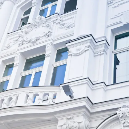 Ein weißes Gebäude mit verzierten weißen Details. Blaue Fenster mit dekorativen Elementen darüber. Ein Balkon mit Geländer.