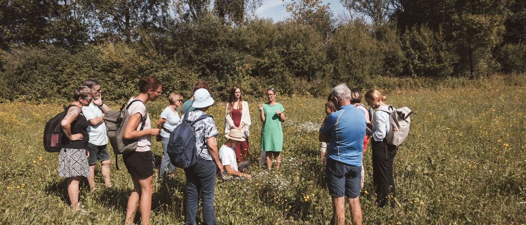 Eine Gruppe von Menschen steht auf einem Feld, einige tragen Hüte und haben Rucksäcke dabei, eine Person kniet und andere schauen sich um, umgeben von hohem Gras und Bäumen.