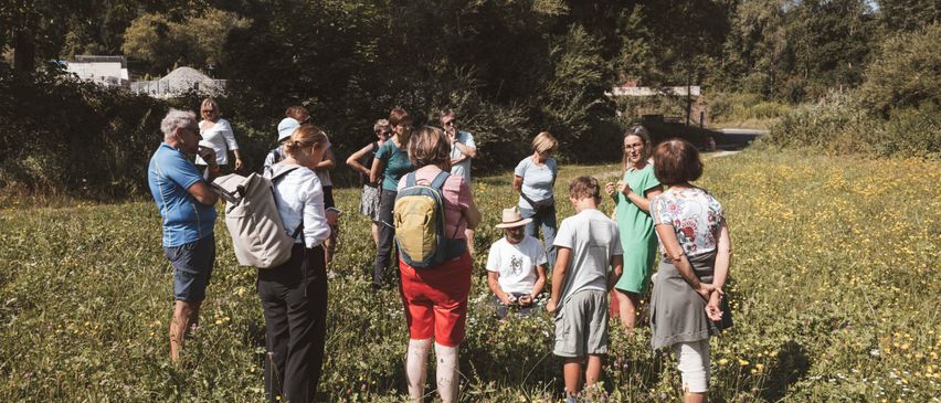 Eine Gruppe von Menschen steht in einer Wiese, einige tragen Hüte und Rucksäcke. Ein Mann sitzt auf dem Boden und beobachtet die Umgebung. Das Gebiet ist von Bäumen umgeben und in der Ferne sind Berge zu sehen.