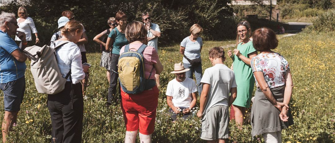 Eine Gruppe von Menschen, einige mit Rucksäcken, steht auf einem Feld. Einige tragen Hüte, Brillen und Sneakers. Eine Person sitzt und hält eine Kamera. Das Feld ist von Bäumen und Pflanzen umgeben.