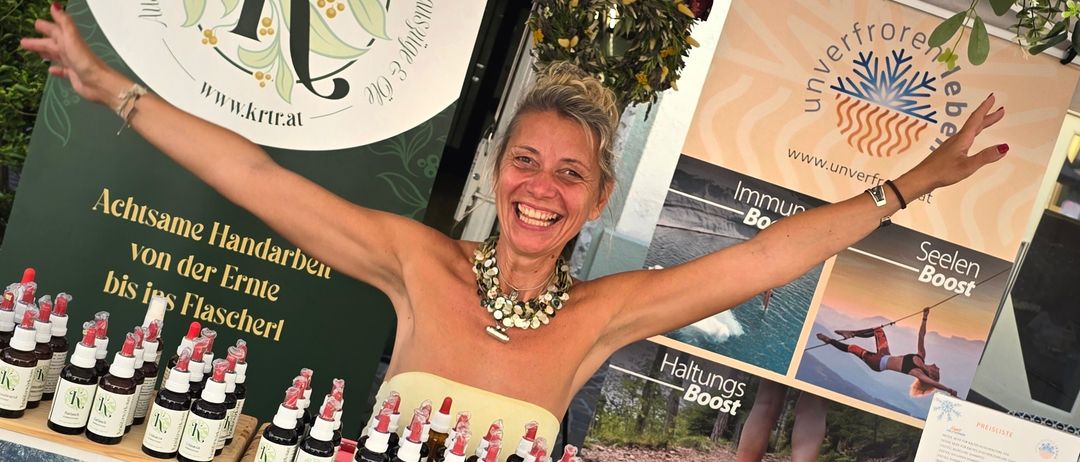 Eine lächelnde Frau steht hinter einem Tisch voller Flaschen in einem Marktstand. Der Stand hat ein Banner mit dem Logo 'Krauterwerkstatt' und deutschem Text. Die Flaschen sind in Reihen angeordnet und mit 'K' beschriftet. Im Hintergrund zeigt ein größeres Banner verschiedene Bilder und Texte.