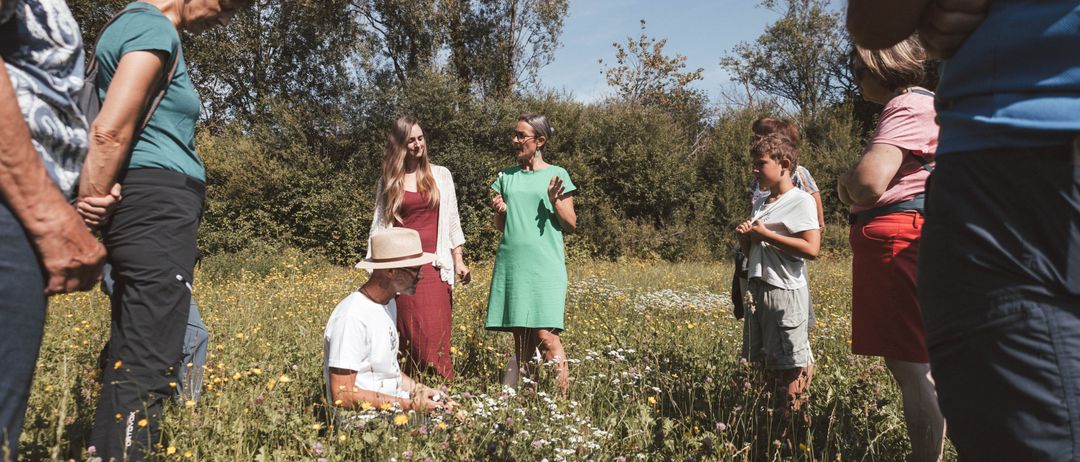 Eine Gruppe von Menschen steht in einem Blumenfeld. Ein Mann sitzt auf dem Boden, umgeben von Blumen. Eine Frau steht mit erhobenen Armen, und eine andere Frau schaut zu ihr. Ein Junge schaut auf den Boden. Bäume umgeben das Feld.