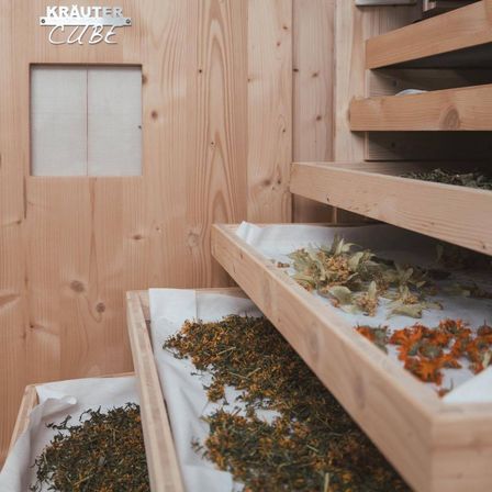 Ein hölzerner Raum mit einem Schild, das 'Krauter Cube' besagt. Es gibt Holzregale mit getrockneten Kräutern darauf. Der Raum hat eine Holztür mit einem kleinen Fenster.