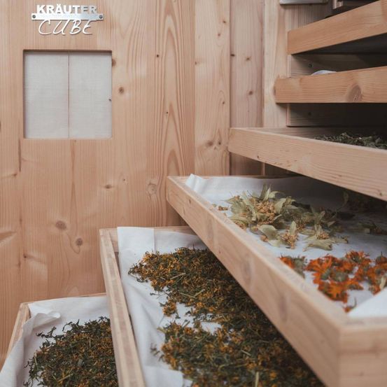 Ein hölzerner Raum mit einem Schild, das 'Krauter Cube' besagt. Es gibt Holzregale mit getrockneten Kräutern darauf. Der Raum hat eine Holztür mit einem kleinen Fenster.