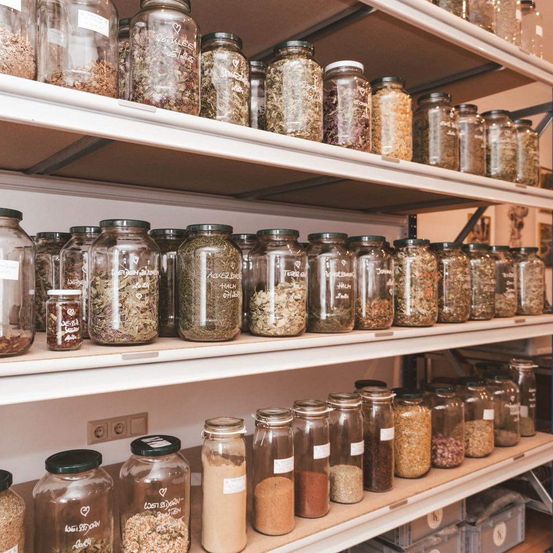 Eine Sammlung von Glasgläsern, gefüllt mit verschiedenen Kräutern und Gewürzen, ist ordentlich auf Regalen angeordnet. Jedes Glas hat einen Etikett mit handgeschriebenem Text. Die Regale sind weiß und ordentlich organisiert.