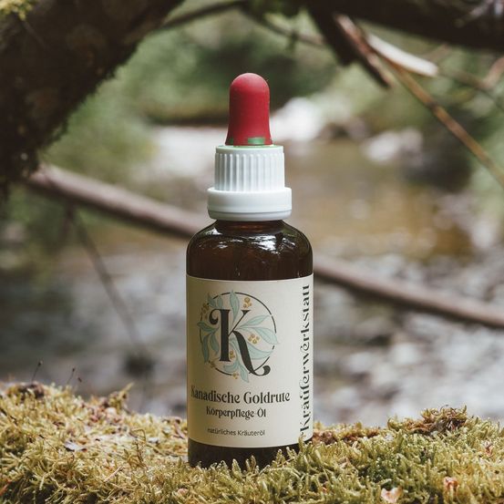 Eine Flasche Kanadische Goldrute Körperpflege Öl, ein natürliches Pflegeöl, ruht auf Moos neben einem Bach. Das Etikett zeigt ein stilisiertes 'K' in einem grünen Kreis.